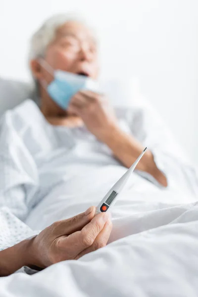 Electronic thermometer in hand on blurred elderly patient in hospital ward — Stock Photo