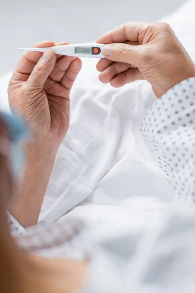 Vista ritagliata del paziente anziano che tiene il termometro elettronico sul letto in clinica — Foto stock