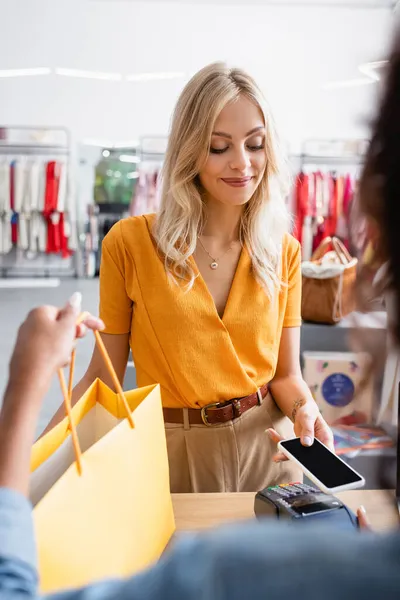 Happy blonde woman paying with smartphone near blurred african american saleswoman in boutique — Stock Photo