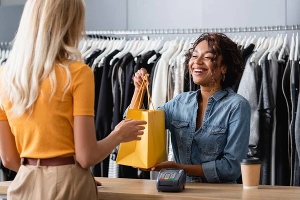 Happy african american saleswoman giving yellow paper bag to client in boutique — Stock Photo