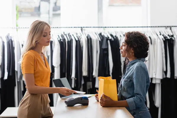 Femme blonde payer avec smartphone près de vendeuse afro-américaine en boutique — Photo de stock