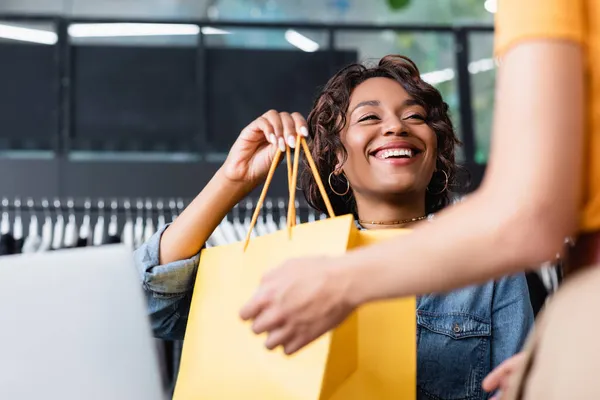 Positive african american saleswoman giving yellow paper bag with purchase to blurred client in boutique — Stock Photo