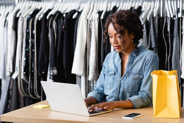 Frisé afro-américaine vendeuse tapant sur le clavier de l'ordinateur portable dans la boutique — Photo de stock