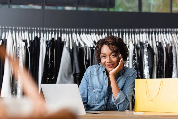 Sonriente afro-americana vendedora mirando cámara en ropa boutique - foto de stock