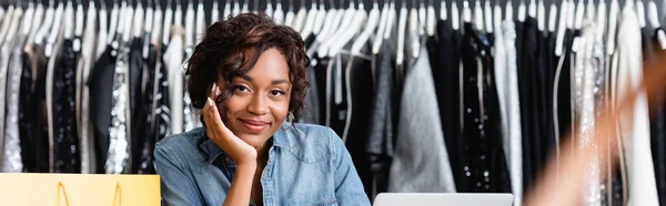 Sonriente afroamericana vendedora mirando a la cámara en la tienda de ropa, pancarta - foto de stock