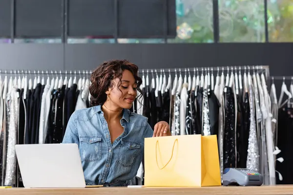 Cheerful african american saleswoman looking at yellow paper bag near gadgets on sales counter desk — Stock Photo