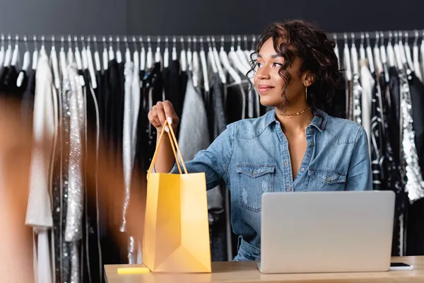 Vendeuse afro-américaine frisée tenant un sac en papier jaune près des gadgets sur le comptoir de vente — Photo de stock