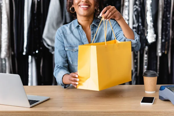 Cropped view of cheerful african american saleswoman holding yellow paper bag near gadgets and paper cup on sales counter desk — Stock Photo