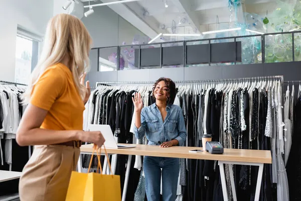 Alegre Africano americano vendedor acenando mão no cliente loiro borrado com saco de compras — Fotografia de Stock