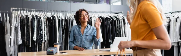 Alegre afroamericana vendedora saludando mano a borrosa rubia cliente con bolsa de la compra, bandera - foto de stock