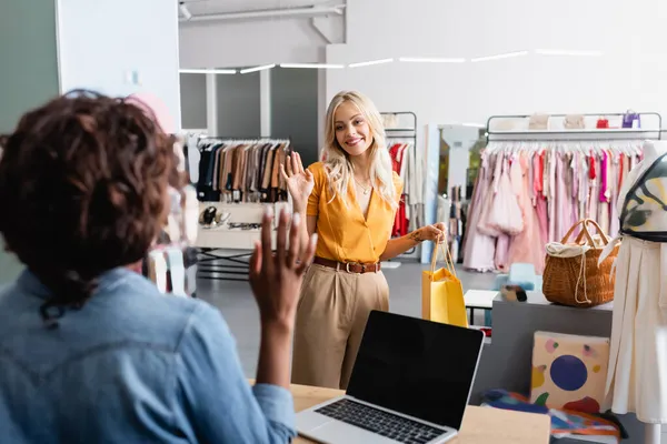 Cliente alegre y rubia con bolsa de la compra agitando la mano a la desenfocada vendedora afroamericana - foto de stock