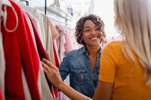 Feliz africano americano tienda asistente mirando rubia cliente en boutique - foto de stock
