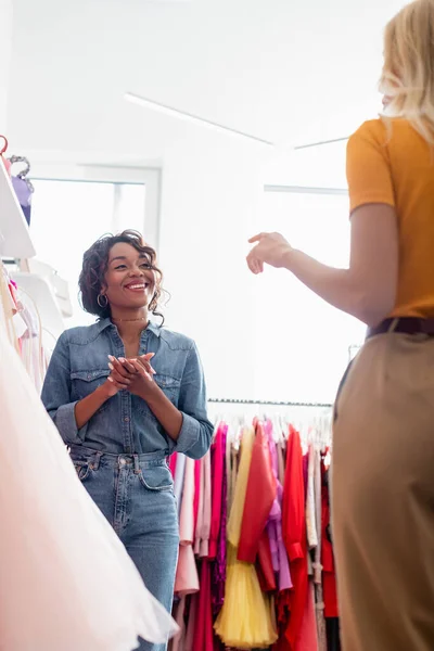 Felice assistente negozio africano americano con le mani serrate guardando il cliente offuscata gesticolando in boutique — Foto stock