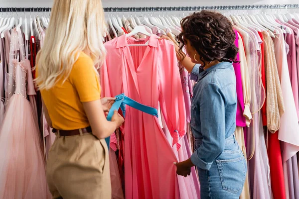 Lockige afrikanisch-amerikanische Verkäuferin zeigt blonder Kundin pinkfarbenes Kleid — Stockfoto