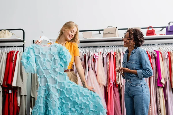 Mulher loira feliz segurando vestido azul no cabide perto de assistente de loja afro-americano na boutique — Fotografia de Stock