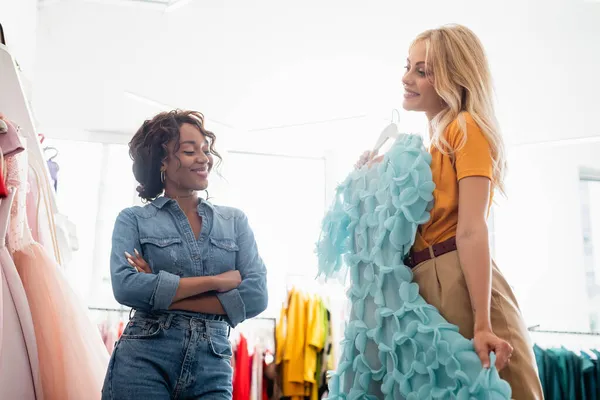 Blonde woman holding blue dress on hanger near smiling african american shop assistant with crossed arms — Stock Photo