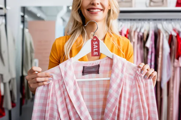Cropped view of cheerful woman holding hanger with checkered pink dress and sale tag — Stock Photo