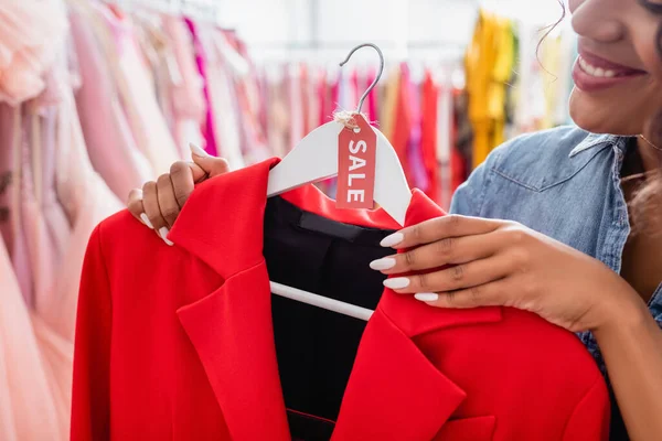 Vista recortada del asistente de la tienda afroamericana feliz celebración chaqueta roja con etiqueta venta en boutique - foto de stock