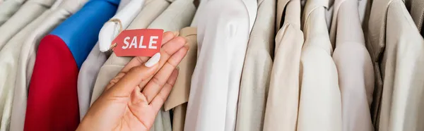 Vista cortada de loja americana africana assistente segurando venda tag perto de roupas em boutique, banner — Fotografia de Stock