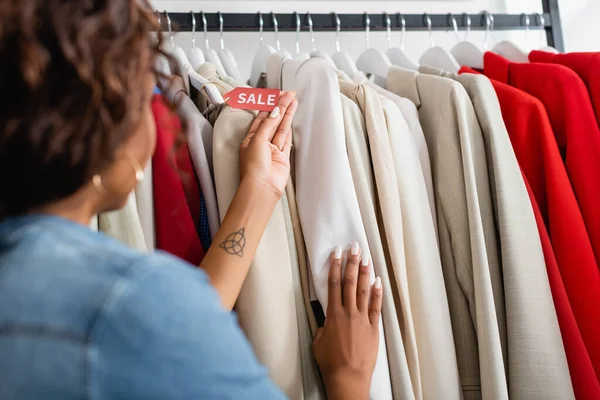 Femme afro-américaine tatouée tenant étiquette de vente près de vêtements en boutique — Photo de stock