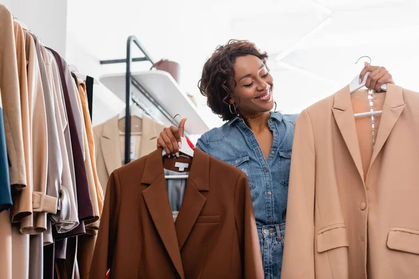 Happy african american shop assistant holding hangers with jackets in boutique — Stock Photo