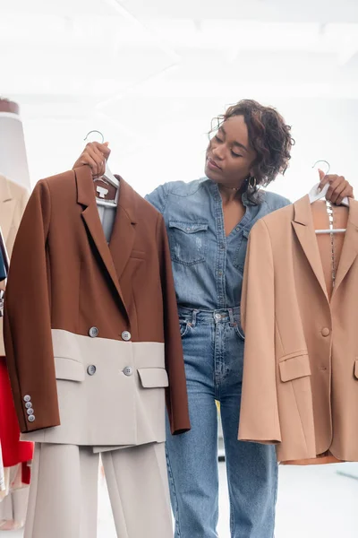 Frisé afro-américain femme tenant cintres avec vestes en boutique — Photo de stock