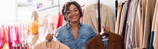 Mulher americana africana alegre segurando cabides com jaquetas em boutique, bandeira — Fotografia de Stock