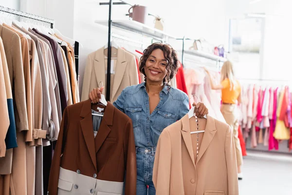 Alegre afroamericana mujer sosteniendo perchas con chaquetas en boutique - foto de stock