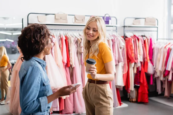 Blonde woman holding paper cup and looking at blurred african american shop assistant with smartphone — Stock Photo