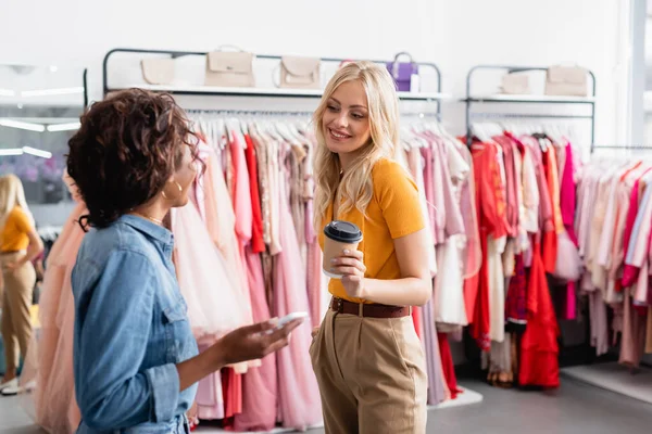 Rubia mujer sosteniendo taza de papel y mirando borrosa africana americana tienda asistente con teléfono inteligente - foto de stock