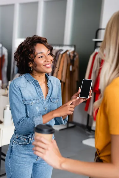 Cheerful african american shop assistant pointing at smartphone with blank screen near blurred customer — Stock Photo