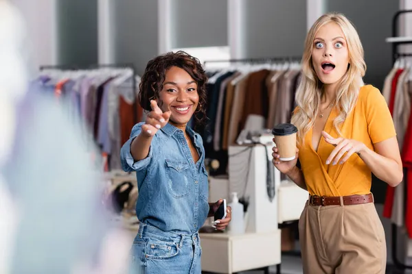 Mulher chocada segurando copo de papel perto de feliz assistente de loja americana africana com smartphone apontando para roupas borradas — Fotografia de Stock