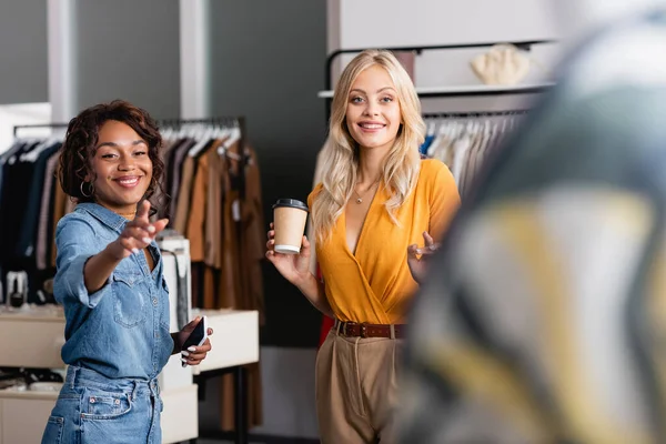 Happy woman holding paper cup while african american shop assistant with smartphone pointing away — Stock Photo
