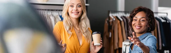 Mujer feliz sosteniendo taza de papel mientras que el asistente de la tienda afroamericana señala con el dedo, bandera - foto de stock