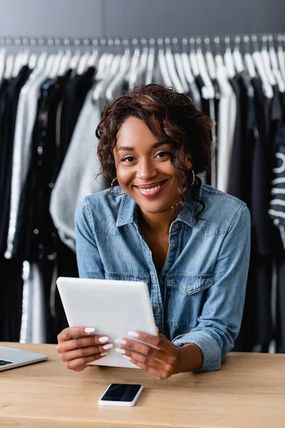 Alegre afroamericana vendedora sosteniendo tableta digital cerca de gadgets en mostrador de ventas - foto de stock