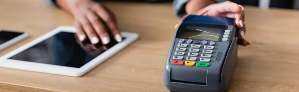 Vista recortada de la vendedora afroamericana sosteniendo terminal de pago cerca de gadgets en mostrador de ventas, pancarta - foto de stock