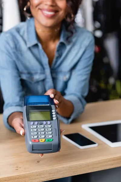 Vista cortada de mulher americana africana feliz segurando terminal de pagamento perto de gadgets no balcão de vendas — Fotografia de Stock