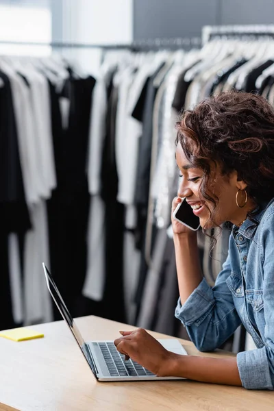 Felice donna afro-americana parlando su smartphone e utilizzando il computer portatile sulla scrivania del banco di vendita — Foto stock