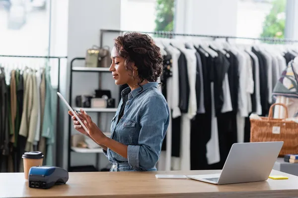 Alegre afroamericana vendedora celebración tableta digital cerca de gadgets y taza de papel en mostrador de ventas - foto de stock