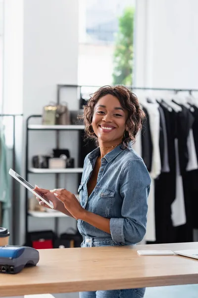 Joyeuse vendeuse afro-américaine tenant tablette numérique près de gadgets et tasse en papier sur le comptoir de vente — Photo de stock