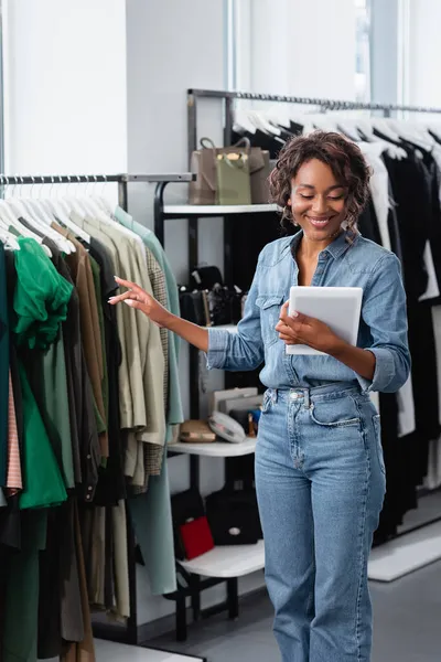 Cheerful african american saleswoman holding digital tablet near clothing on rack — Stock Photo