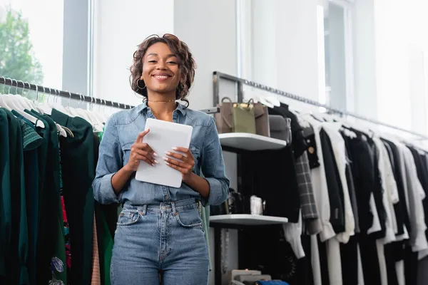 Vendedora afroamericana positiva sosteniendo tableta digital cerca de la ropa en rack - foto de stock