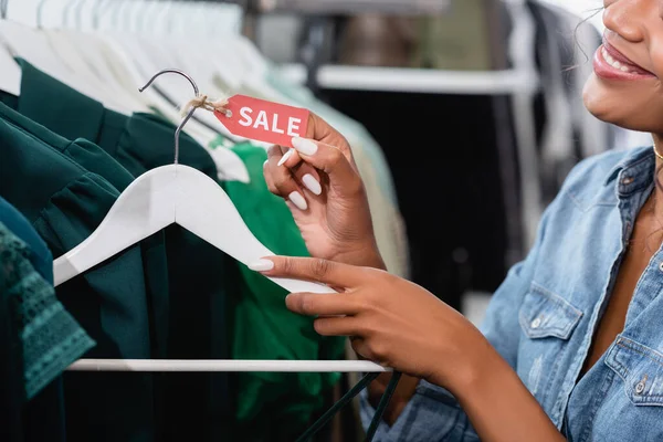 Vista cortada da vendedora americana africana alegre segurando cabide com etiqueta de venda em boutique de roupas — Fotografia de Stock