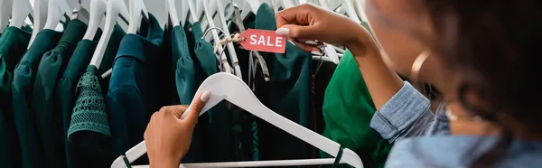 Обрезанный вид на африканскую американскую продавщицу держа вешалку с биркой продажи в магазине одежды, баннер — стоковое фото