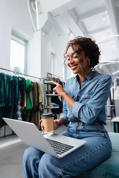 Happy african american woman pointing with finger during video chat in clothing boutique — Stock Photo