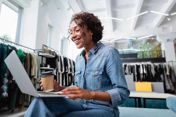 Mujer afroamericana feliz mirando el ordenador portátil y sosteniendo la taza de papel en la boutique de ropa - foto de stock