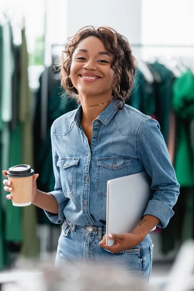 Gaie femme afro-américaine tenant ordinateur portable et tasse en papier dans la boutique — Photo de stock