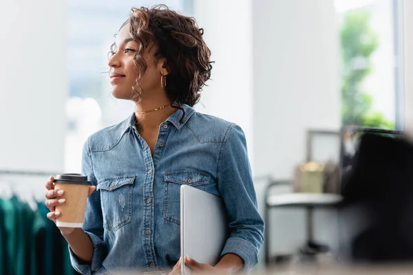 Piacere afroamericano donna in possesso di computer portatile e tazza di carta in boutique — Foto stock