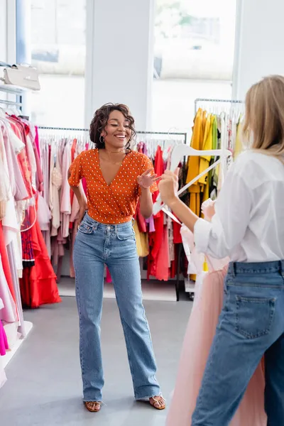 Happy african american woman looking at blonde friend holding pink dress on hanger — Stock Photo