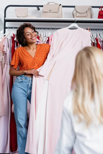 Sorrindo afro-americano mulher segurando cabide com vestido rosa perto de amigo loiro — Fotografia de Stock